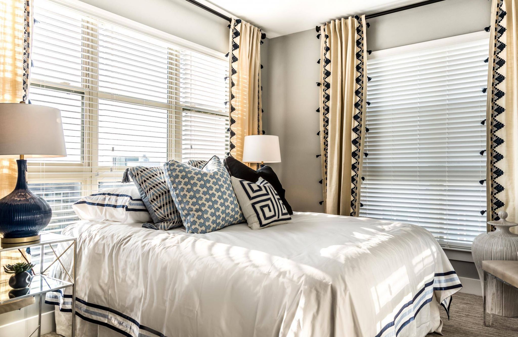Bright and stylish bedroom at Carolina Square featuring neutral bedding, patterned accent pillows, elegant drapery, and faux wood blinds on large windows.