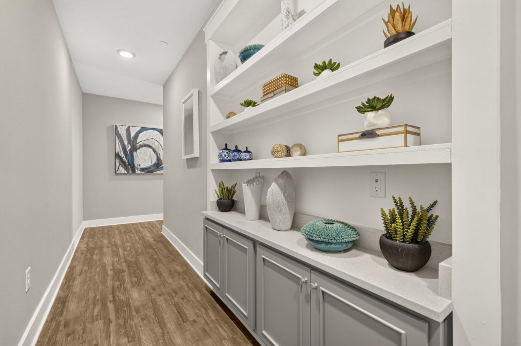 A hallway with wood floors, built-in white shelves displaying vases, plants, and decor, and abstract art on the wall at the end.