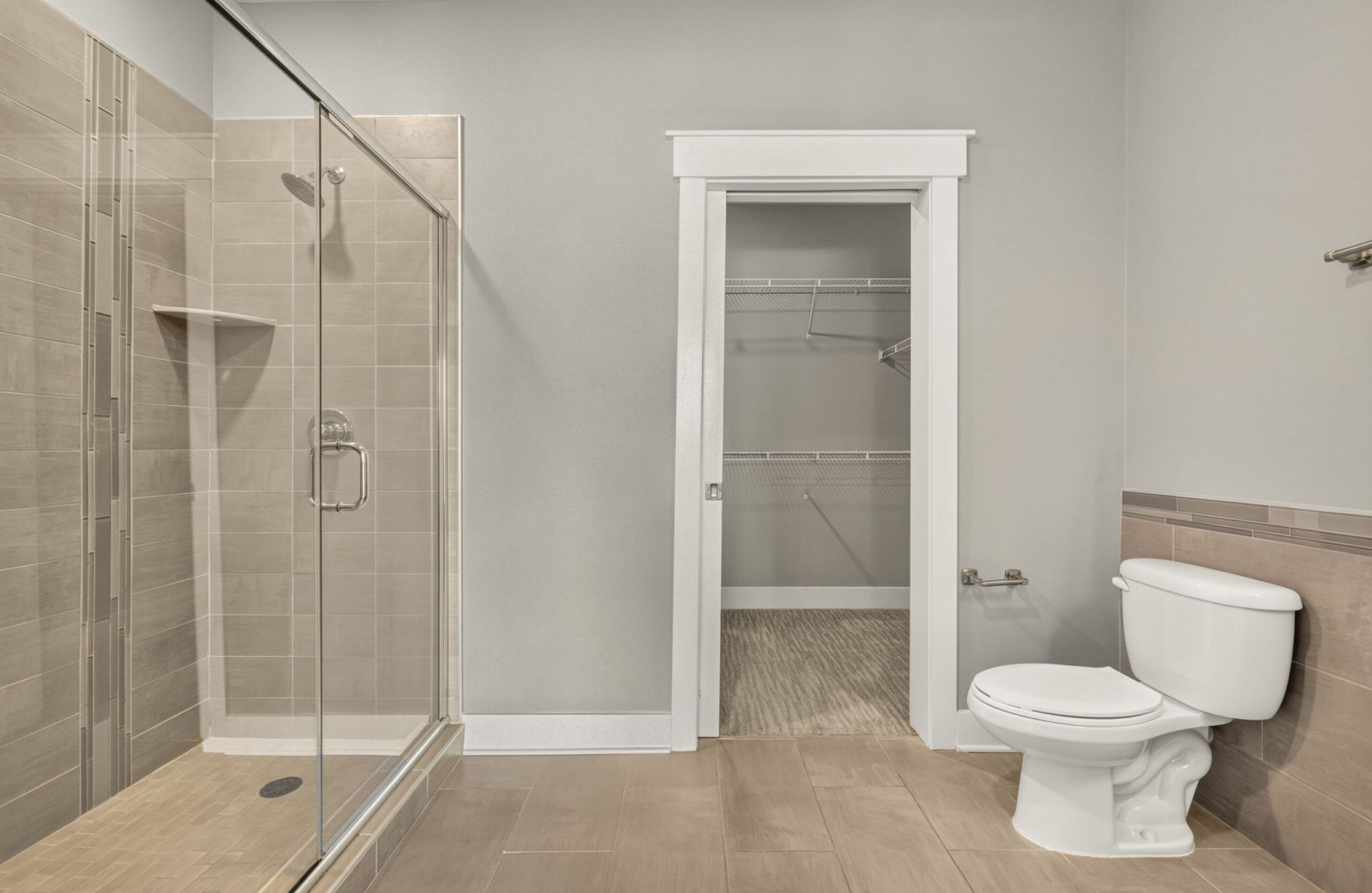Modern bathroom with glass shower, toilet, tan tile floor, and open door leading to a walk-in closet with shelves.