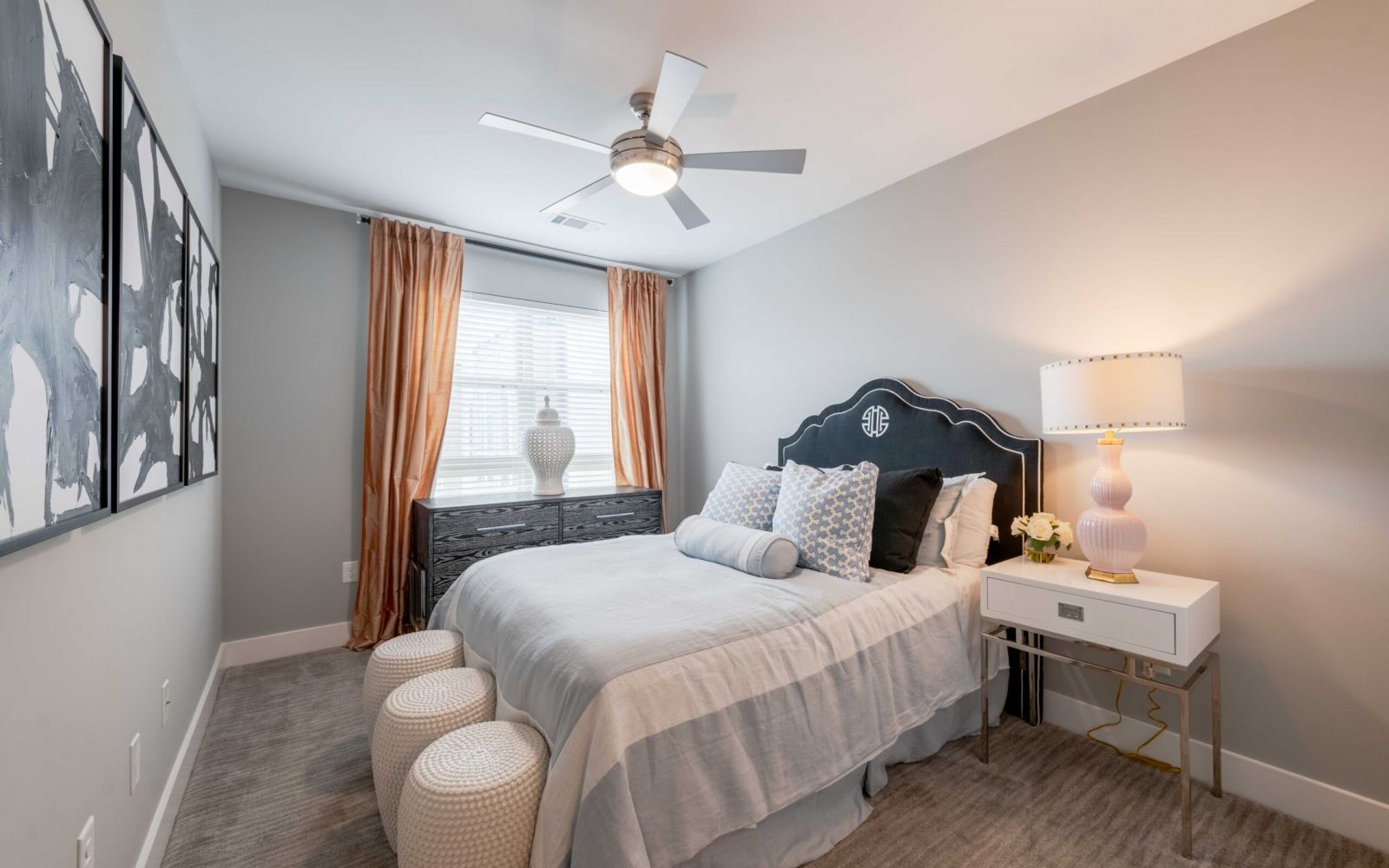 Modern bedroom with a bed, two stools, dresser, lamp, and large window with curtains letting in daylight.
