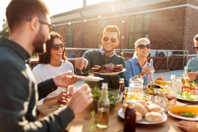 Four friends wearing sunglasses enjoy food and drinks together at an outdoor table on a sunny day.
