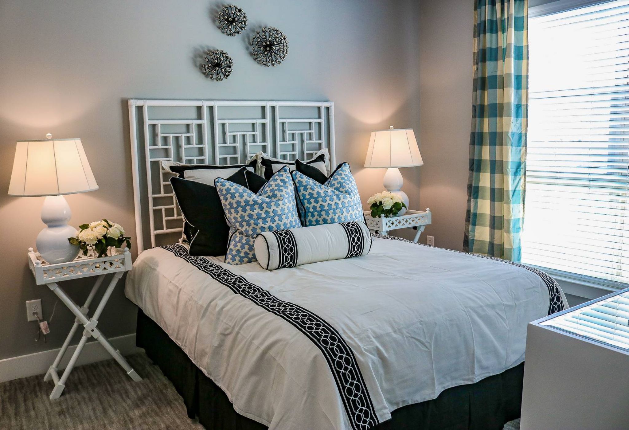 Modern bedroom with white bed, blue accents, two lamps, floral decor, and plaid curtains by a sunny window.