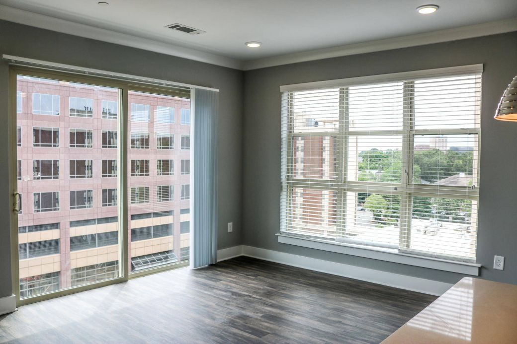 Modern apartment room with large windows, blinds, and wood floors, overlooking city buildings and greenery at Carolina Square apartments