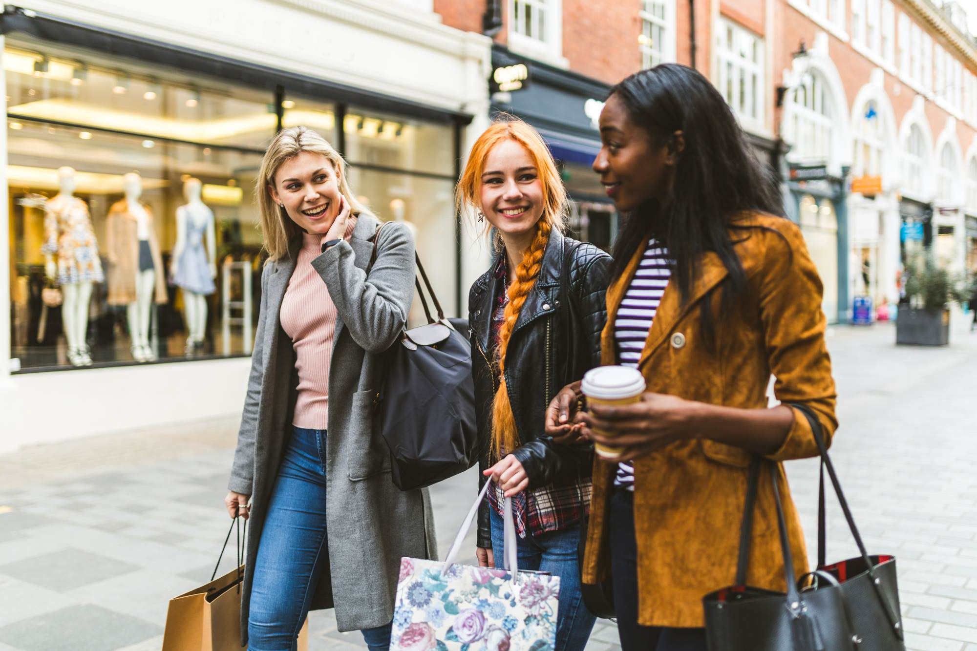 Three young women smiling and walking outside shops, carrying shopping bags and a coffee cup.