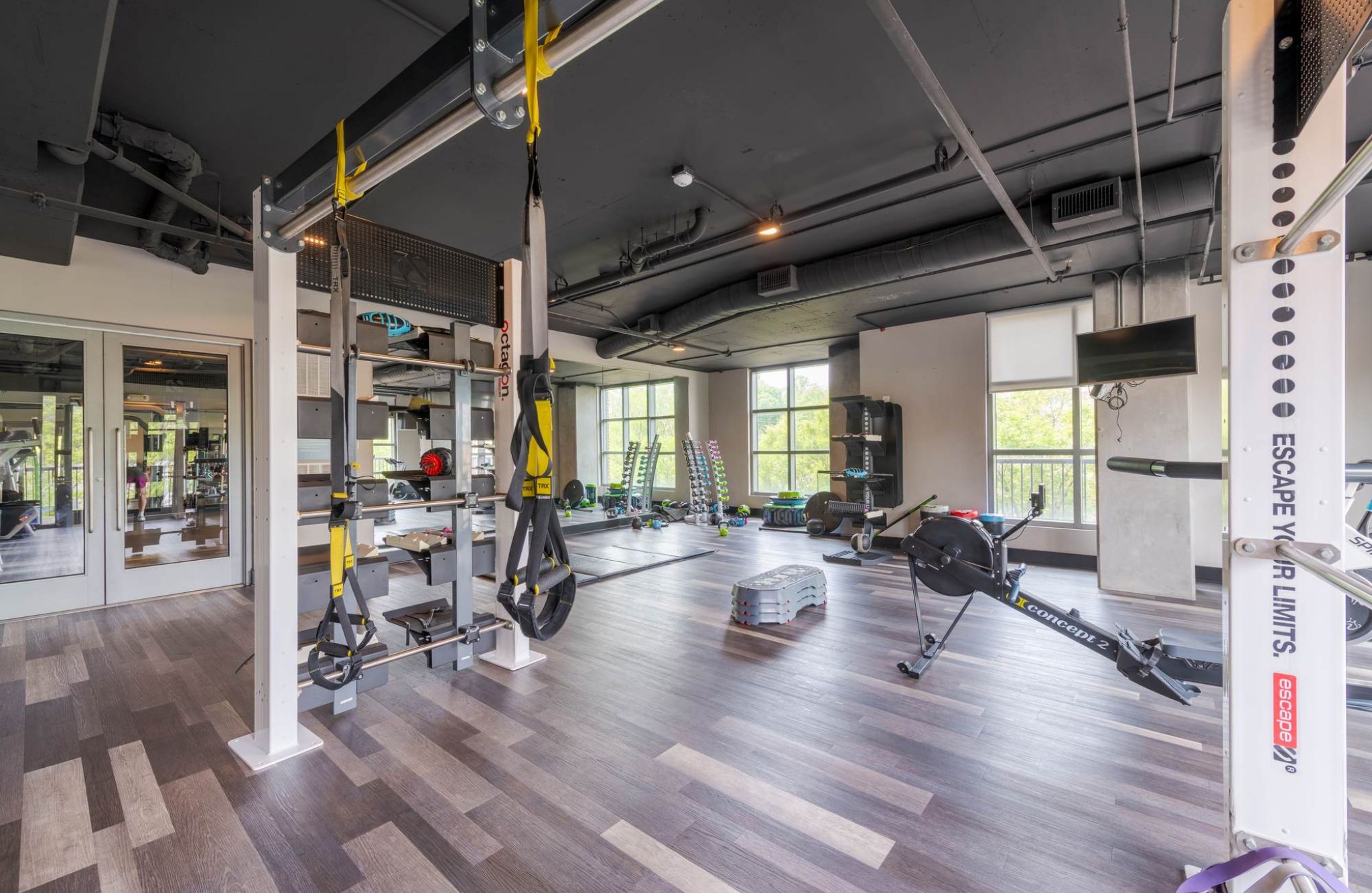 Modern gym with exercise equipment, large windows, wood flooring, and various workout machines and weights.