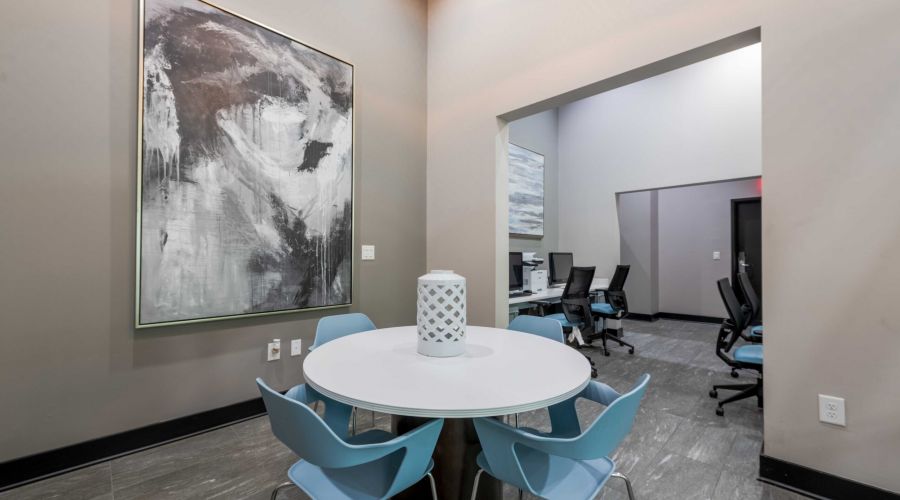 Round table with four blue chairs, large abstract painting, and office workspace in the background.