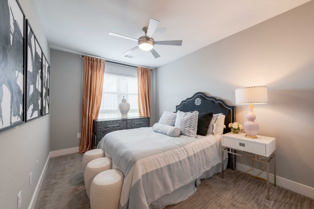 Modern bedroom with a bed, two stools, dresser, lamp, and large window with curtains letting in daylight.