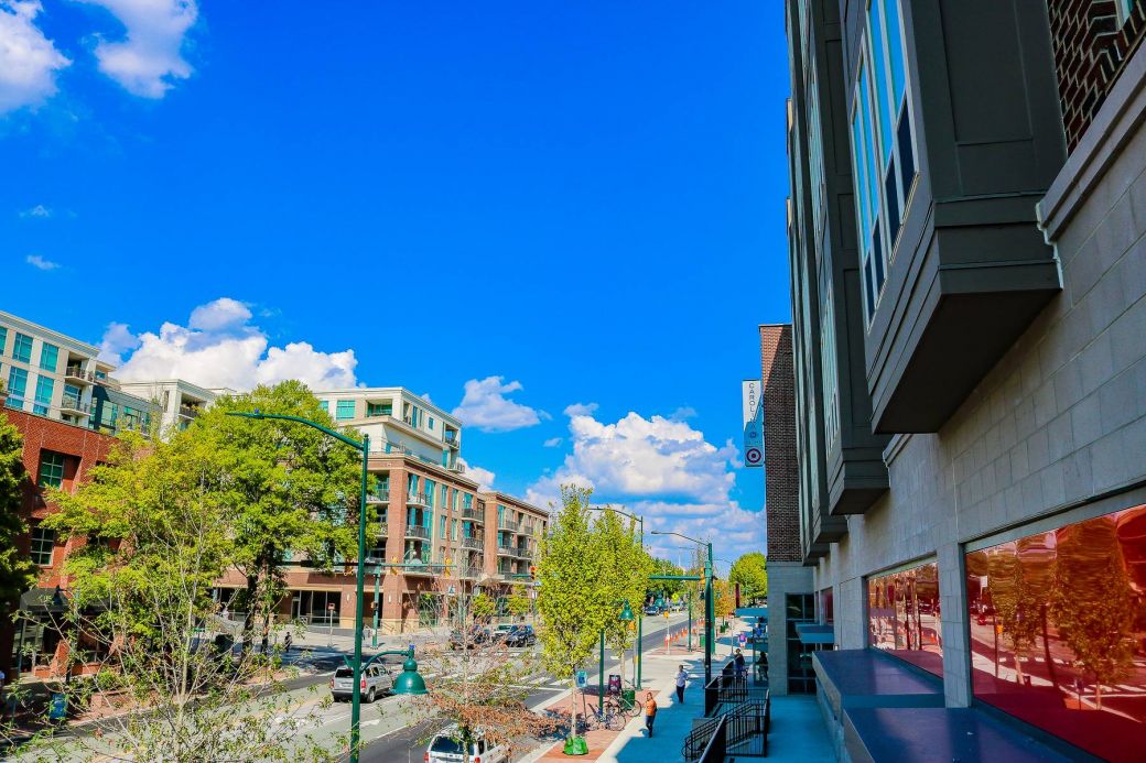 A sunny city street lined with modern buildings, trees, and parked cars under a bright blue sky.