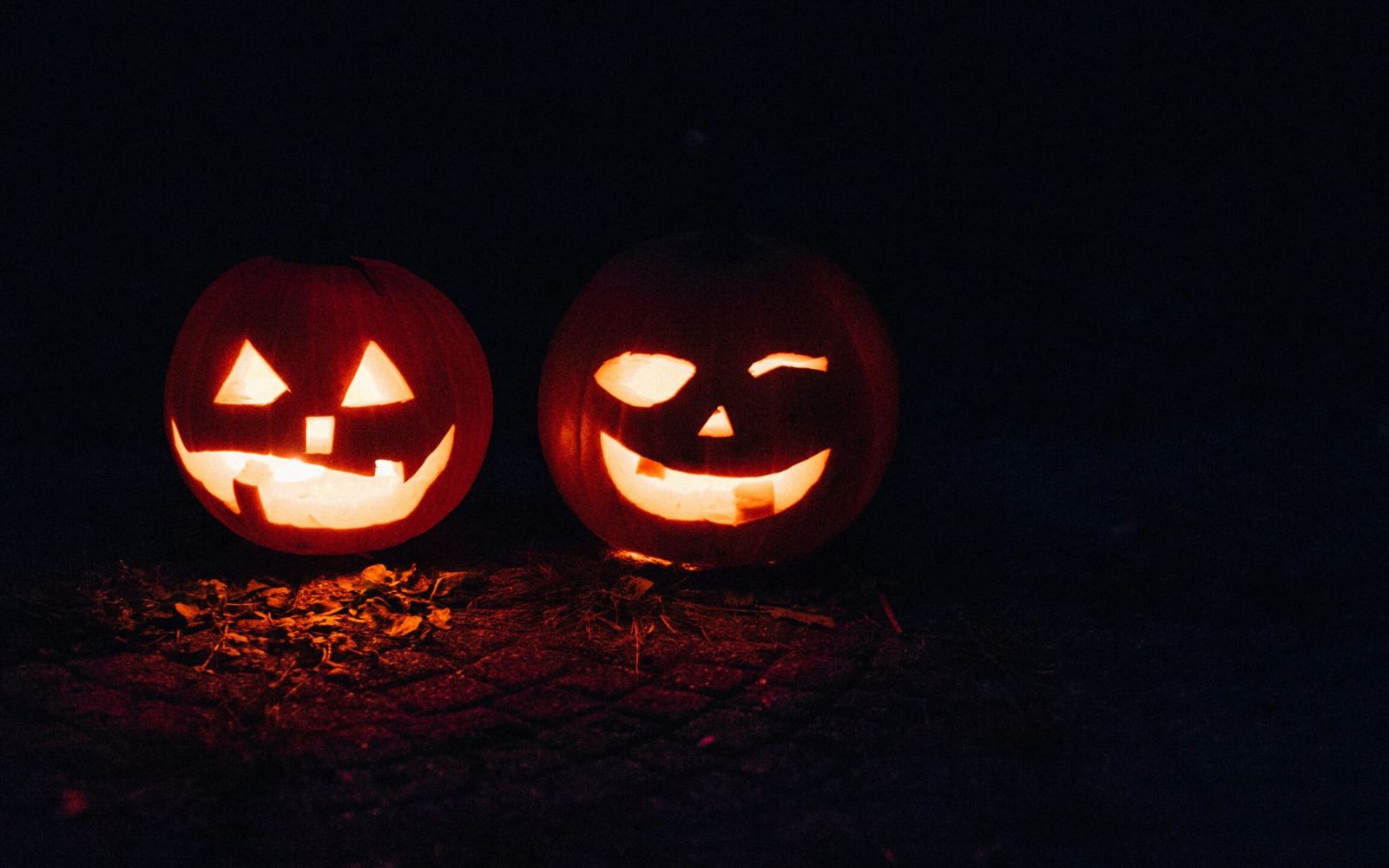 Two glowing jack-o’-lanterns with carved faces sit side by side on the ground in the dark.