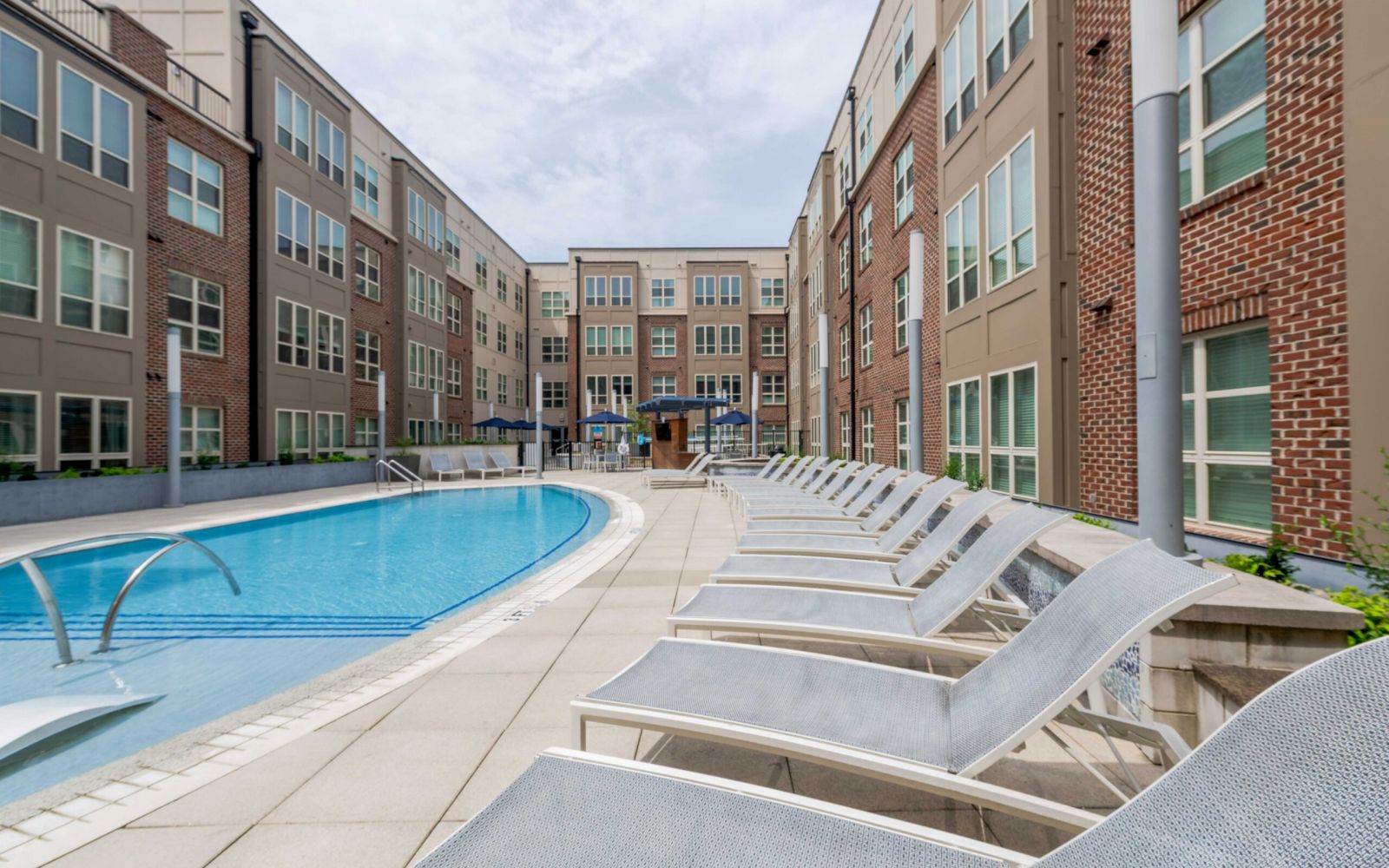 Lounge chairs line a curved outdoor pool next to a modern apartment building under a cloudy sky.