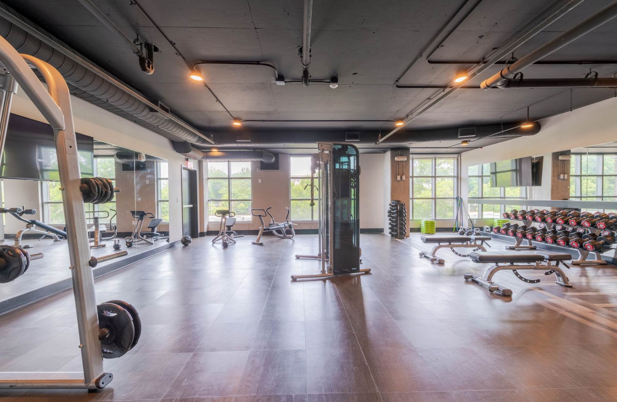 Spacious gym with weight racks, benches, exercise bikes, and large windows letting in natural light.