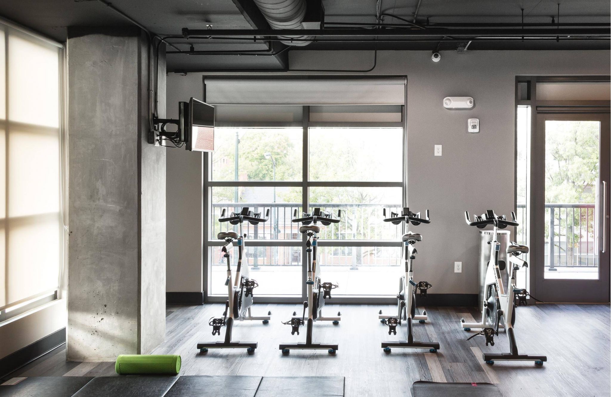 Three stationary bikes in a modern gym with large windows and a green foam roller on the floor.