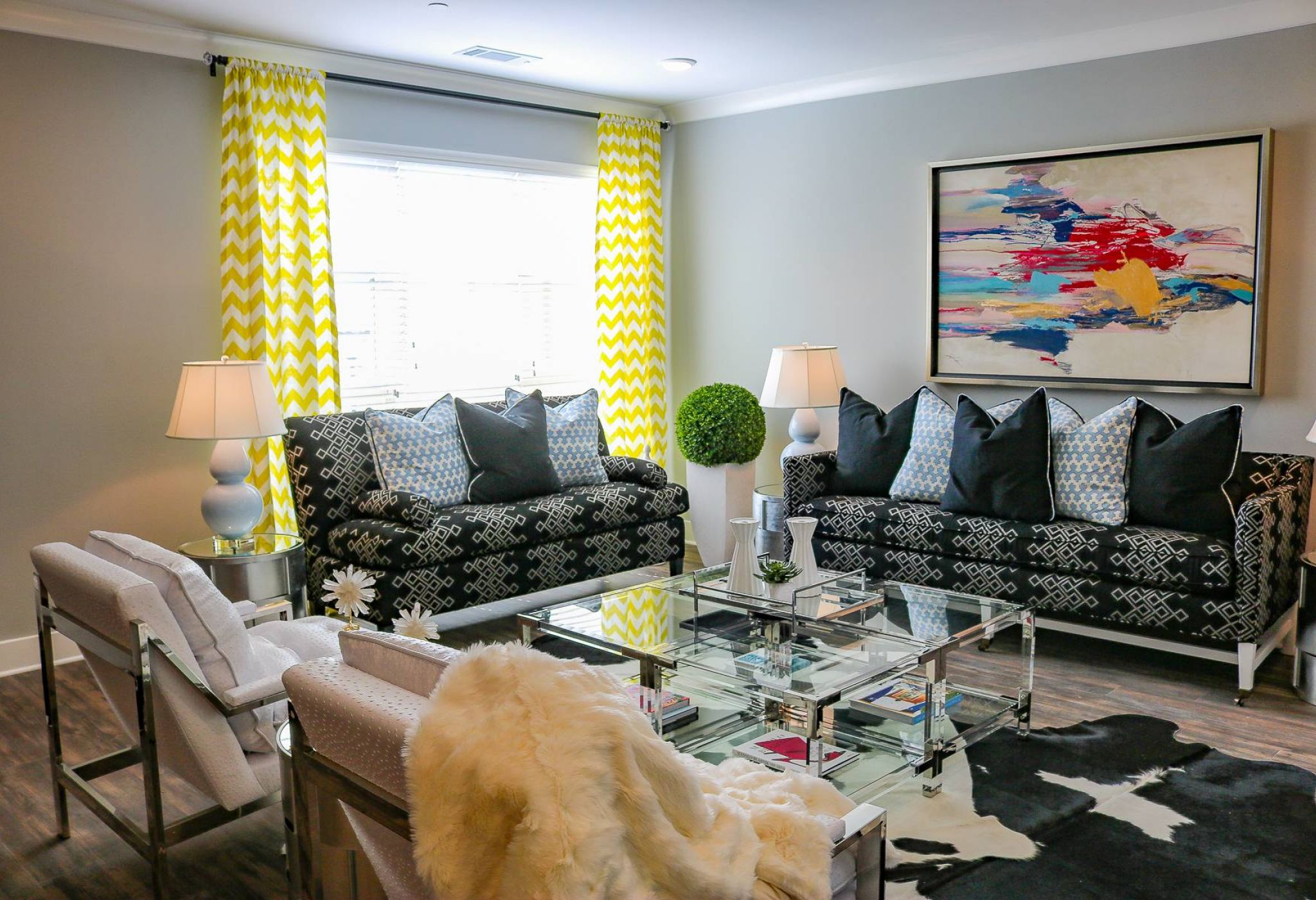 Modern living room with patterned sofas, yellow curtains, abstract art, and glass coffee table.