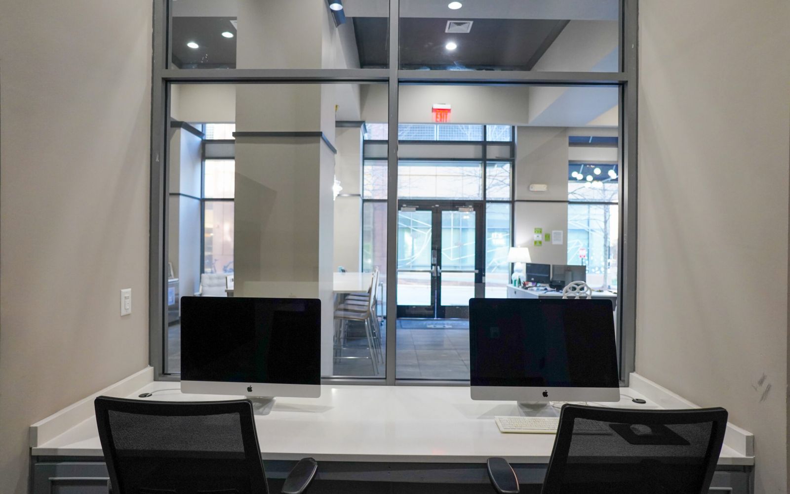 Two desktop computers on a desk in front of large windows inside a modern office space.