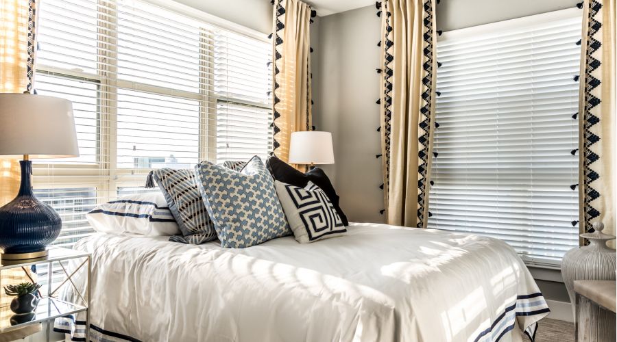 Bright bedroom with large windows, white bedding, patterned pillows, and beige curtains with black trim.