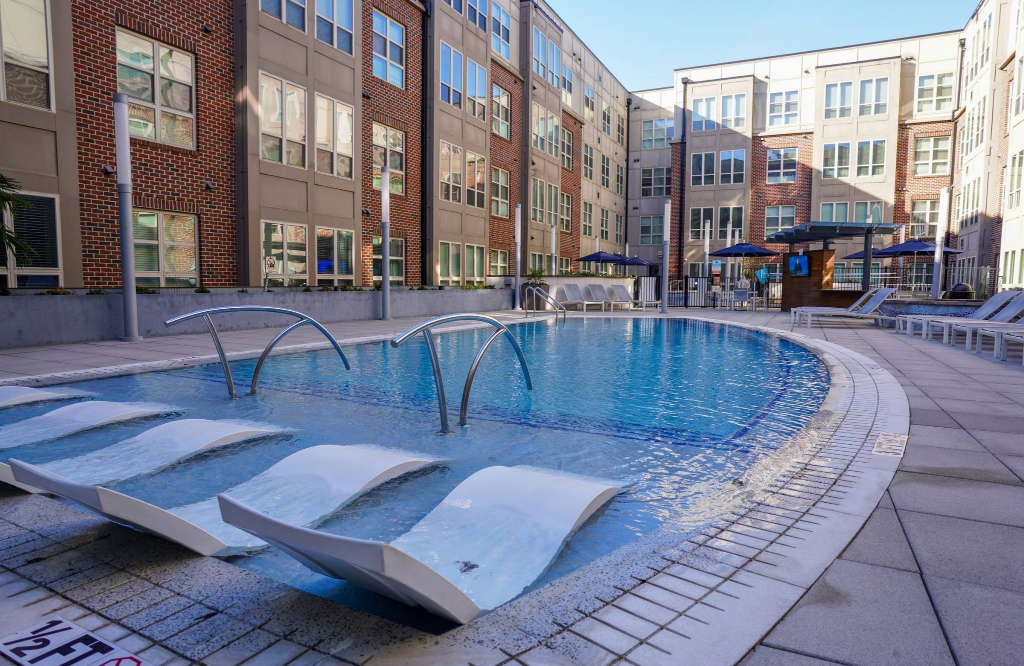 Outdoor apartment pool with lounge chairs, curved metal water features, and surrounding modern brick buildings.