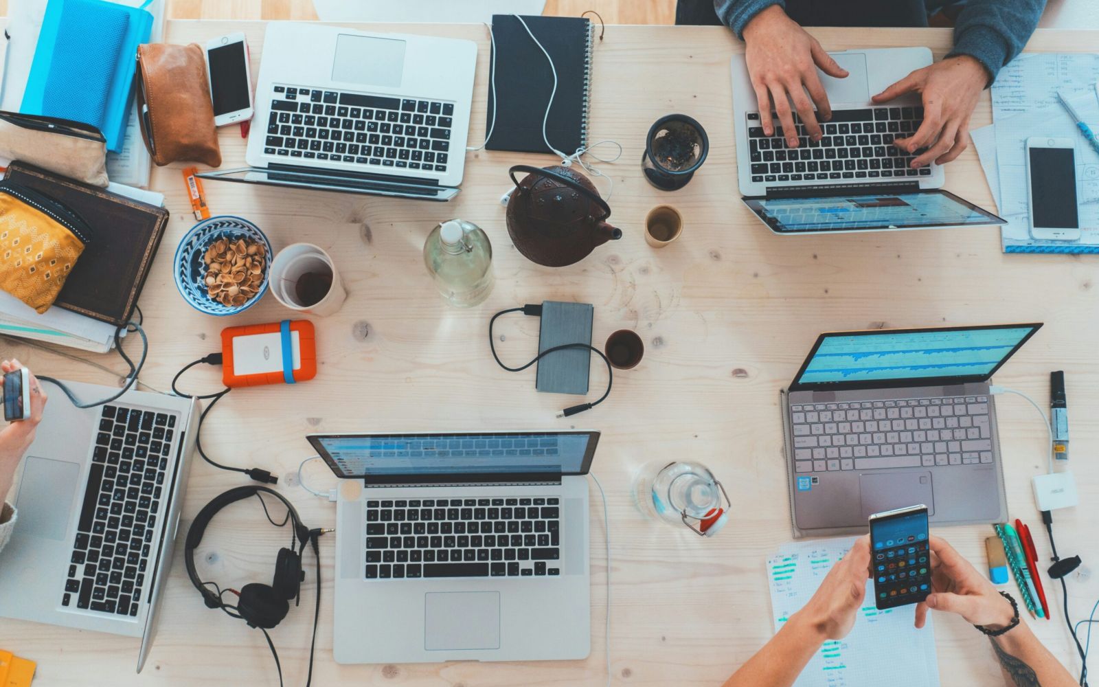 Overhead view of a cluttered work desk with laptops, notebooks, snacks, and people working together.