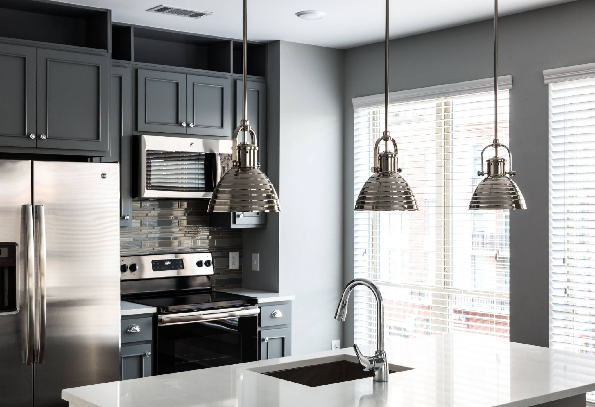 Modern kitchen with stainless steel appliances, gray cabinets, pendant lights, and large windows with blinds.
