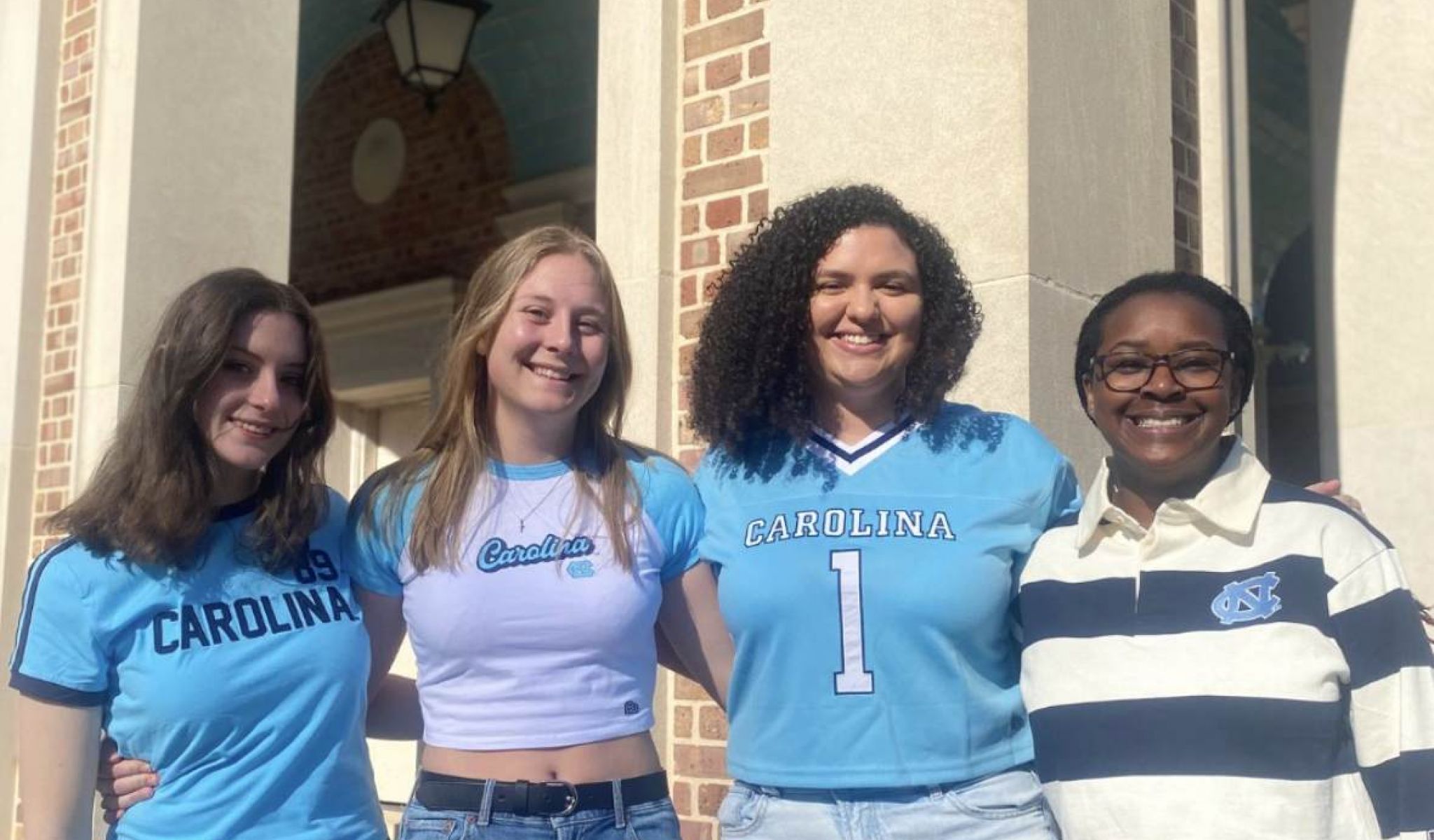 Four young women in UNC Chapel Hill shirts stand smiling together outside a brick building.