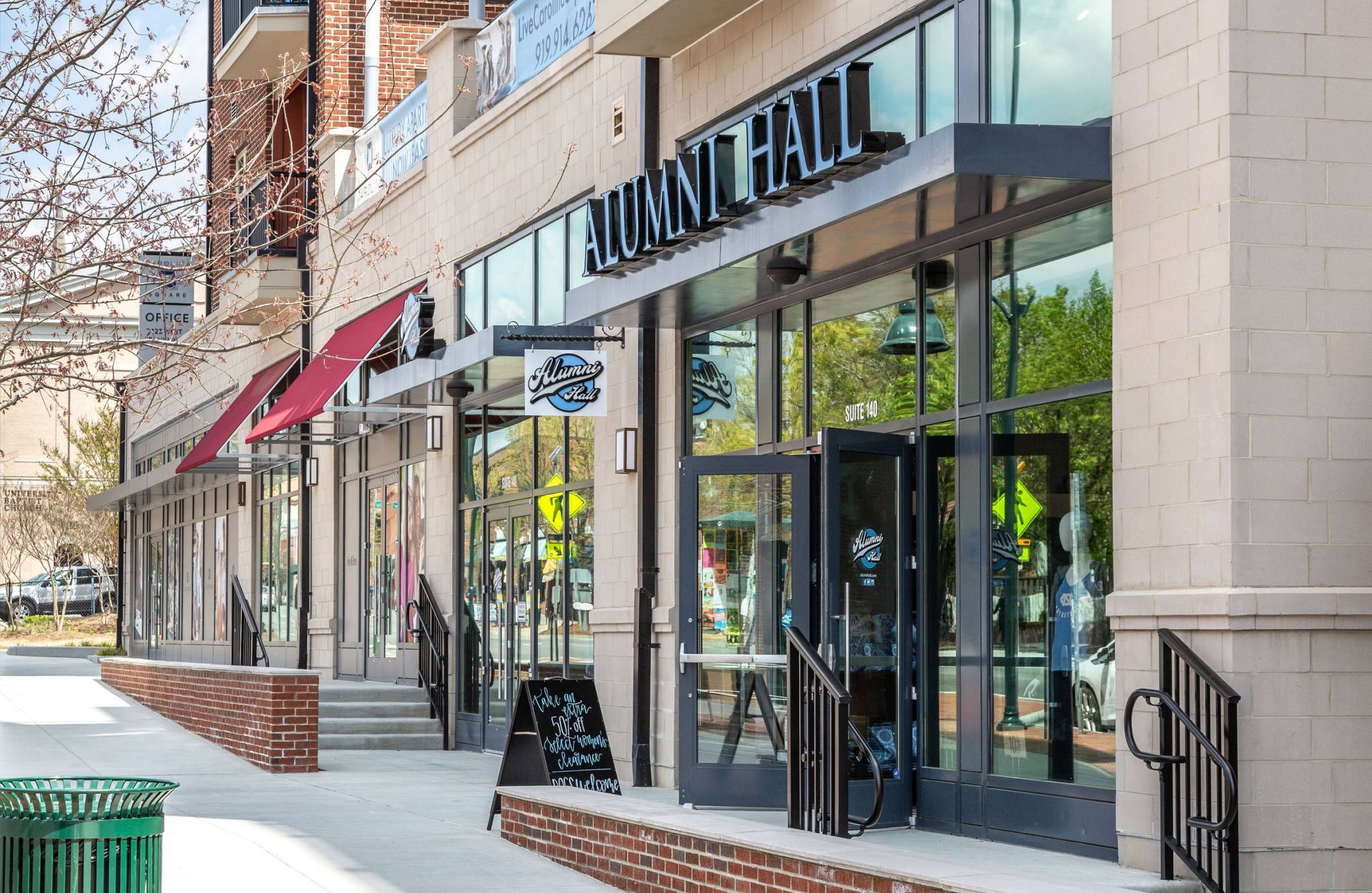 Storefronts with glass windows, including "Alumni Hall," line a sunny sidewalk in a commercial area.