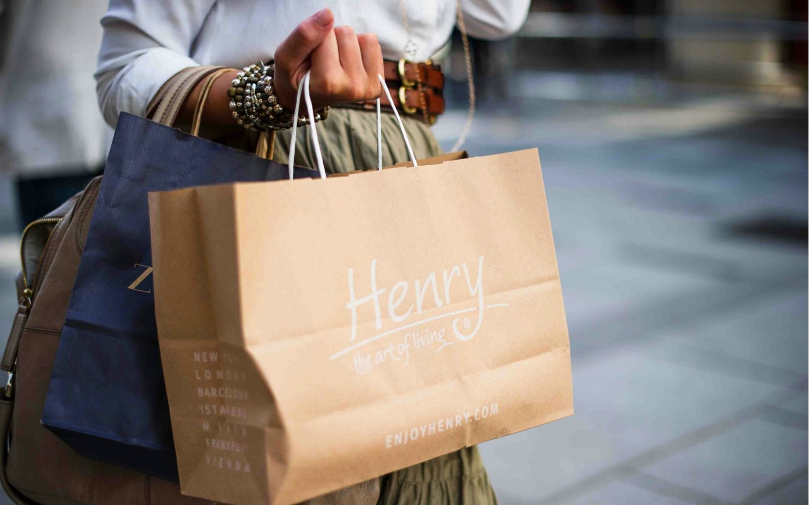 Person holding shopping bags, including a brown one labeled “Henry the art of living,” on a city street.