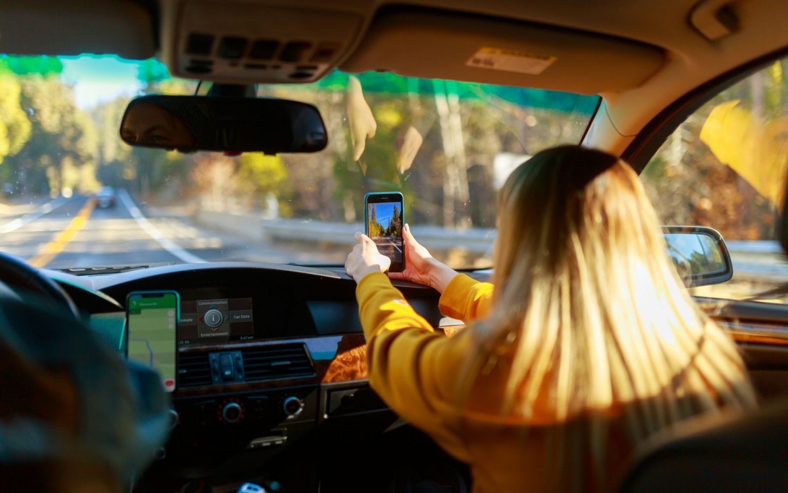 A woman in a car takes a photo with her phone while riding on a sunny road through a forest.