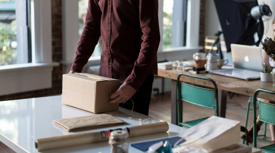 Person in a maroon shirt packing a cardboard box on a white desk with office supplies nearby.