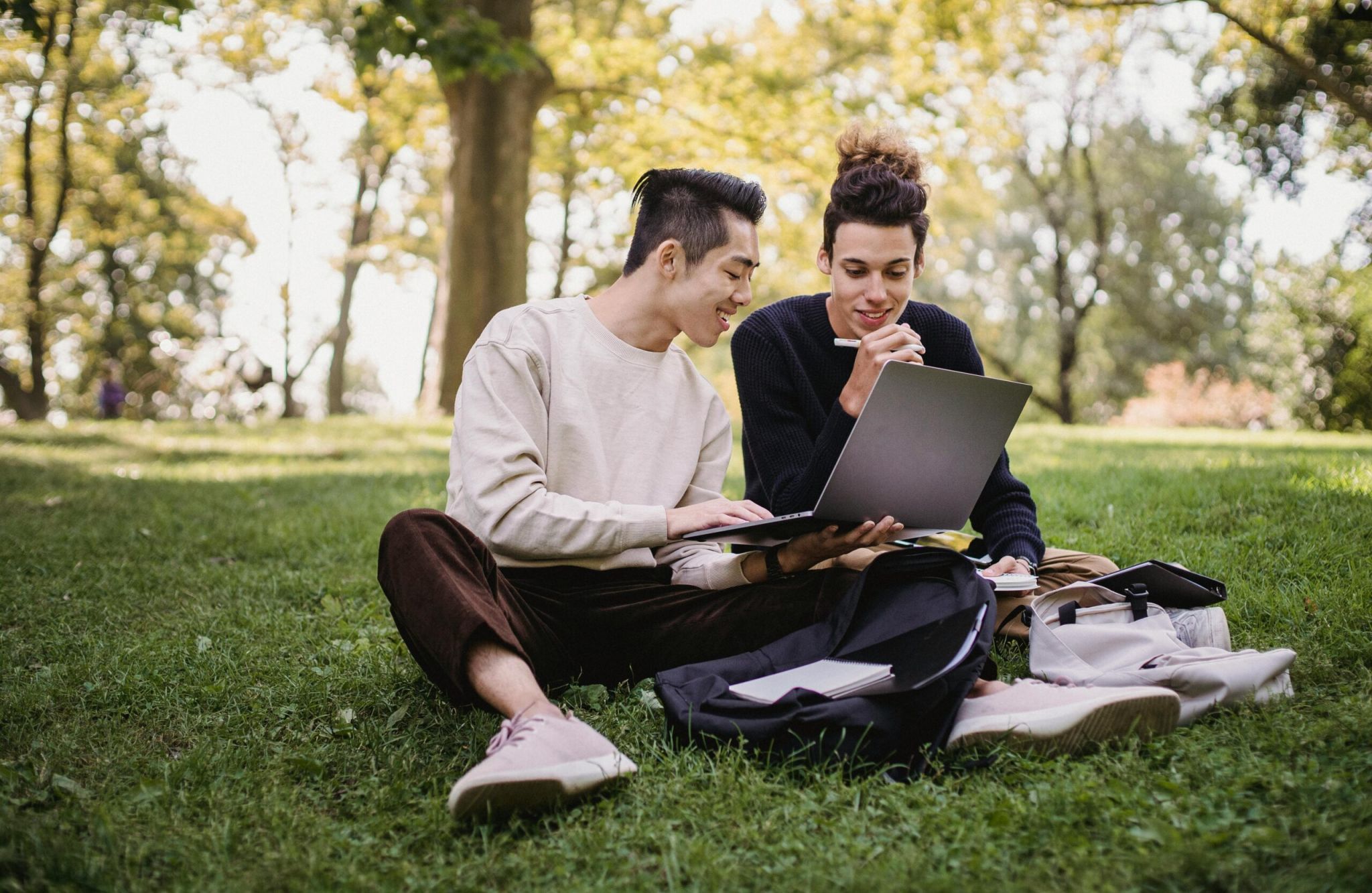 Two people sitting on grass in a park, smiling and working together on a laptop with notebooks nearby.