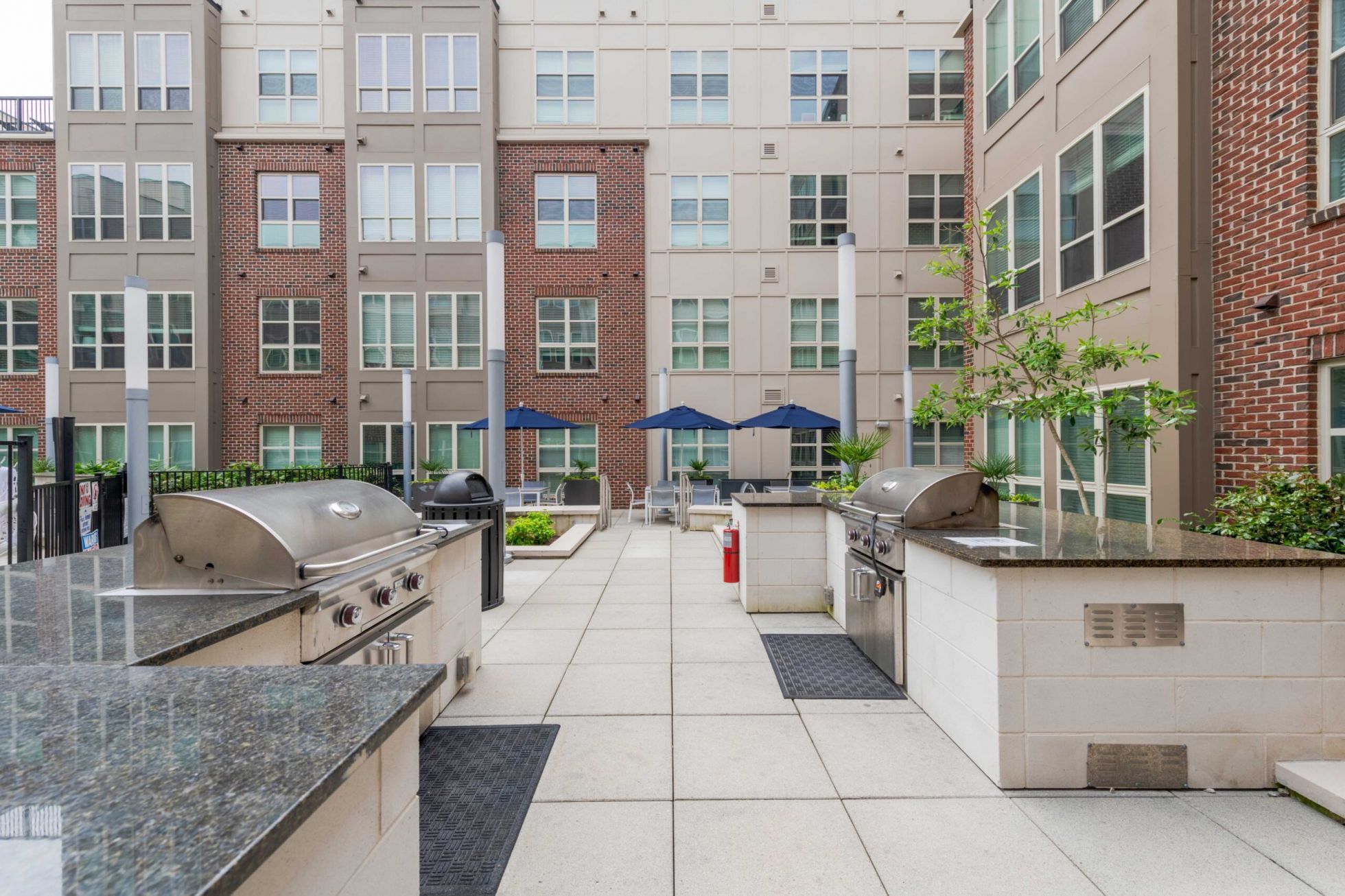 Outdoor apartment courtyard with grilling stations, seating area, and blue umbrellas on a paved patio.
