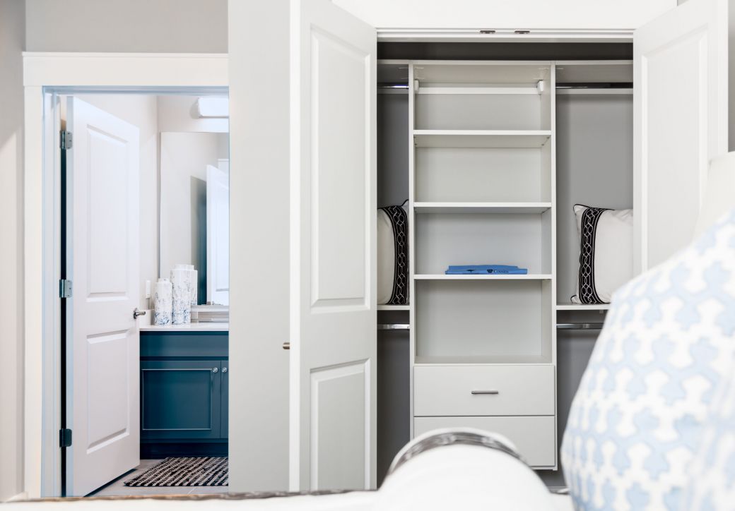 Open closet with white shelves and pillows, next to a bathroom with a blue vanity and open door.
