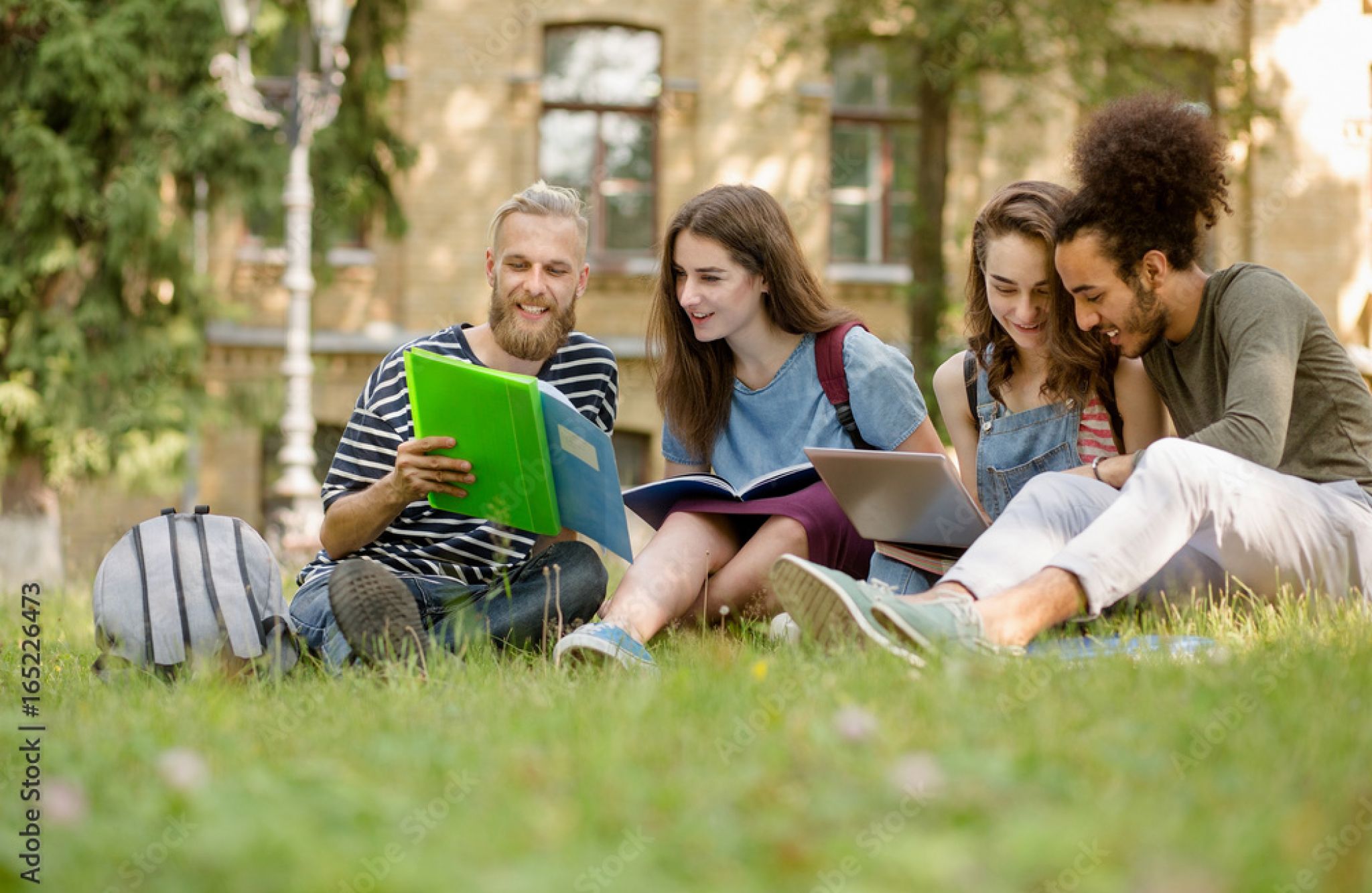 Four students sitting on grass outside, studying together with books and a laptop, smiling and engaged.