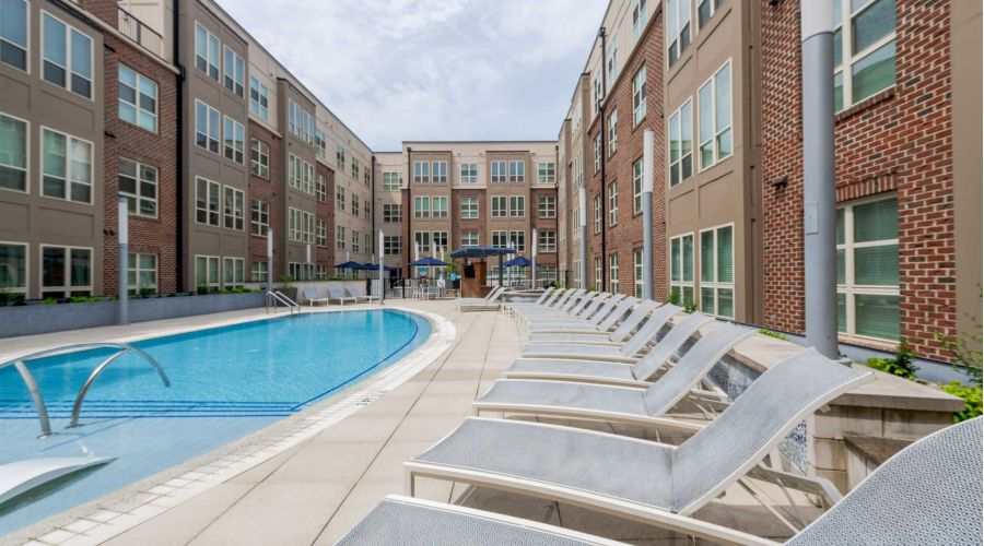 Lounge chairs line a curved outdoor pool next to a modern apartment building under a cloudy sky.