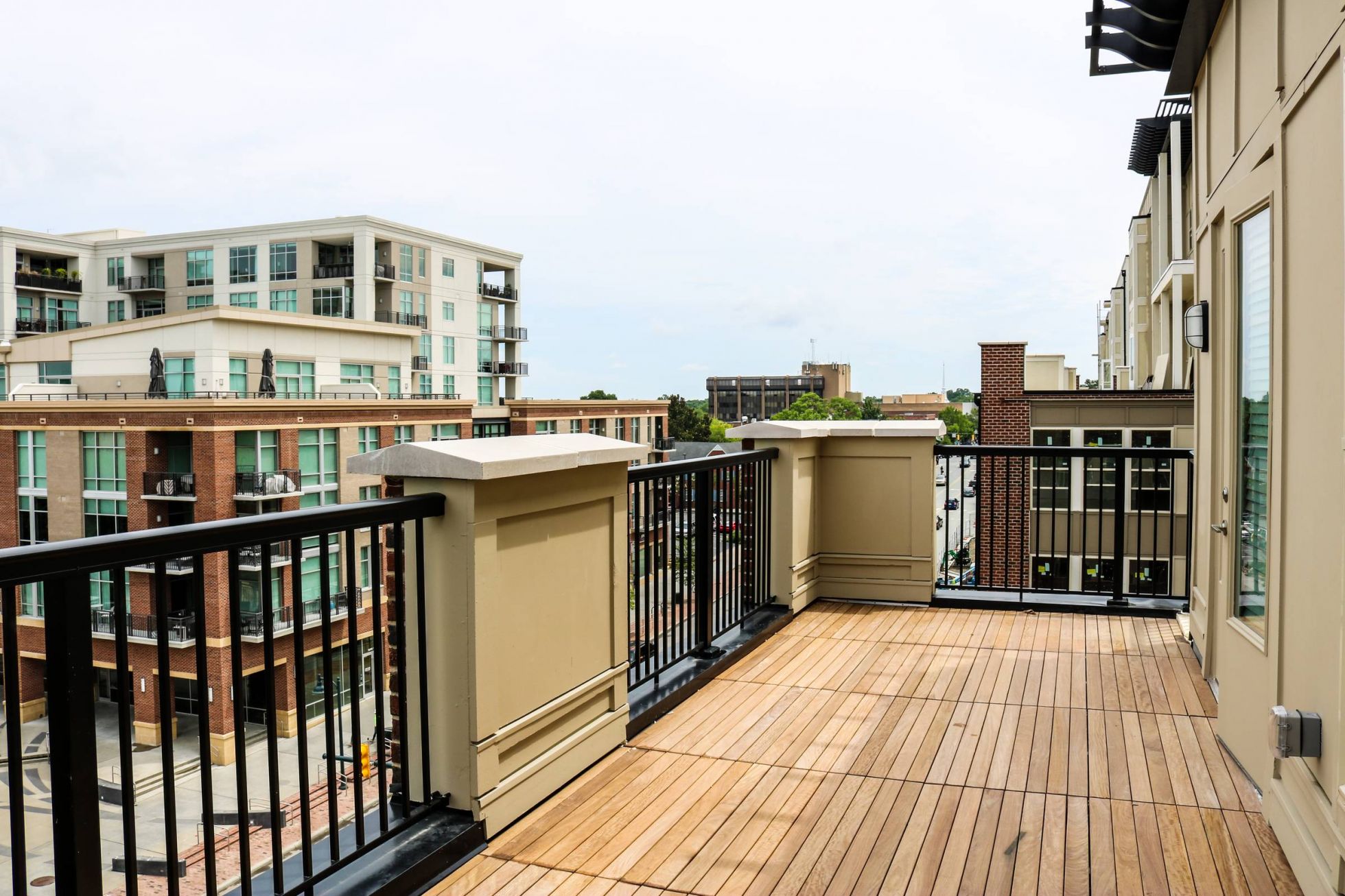 Modern apartment balcony with wood flooring and black railings overlooking nearby buildings and street at Carolina Square Apartments.