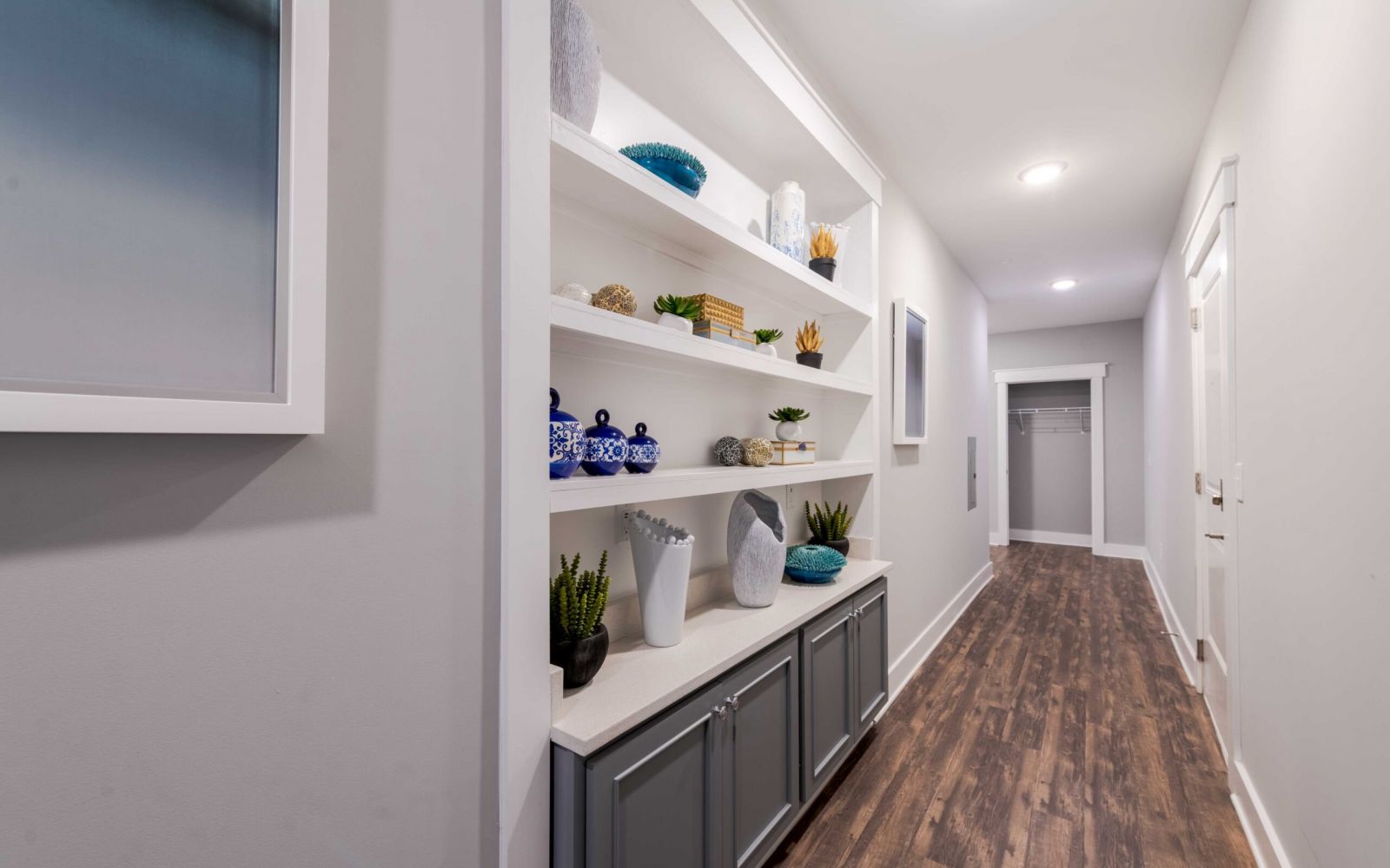 A hallway with wooden floors, built-in shelves holding decor, and an open closet at the end.