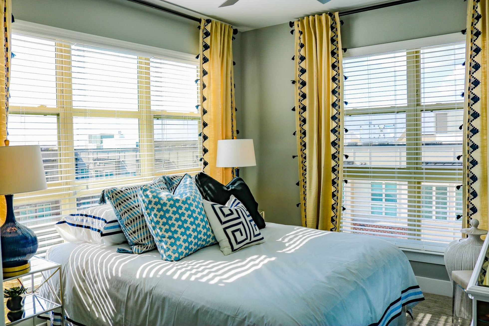 Bright bedroom with yellow curtains, patterned pillows, and sunlight streaming through large windows.
