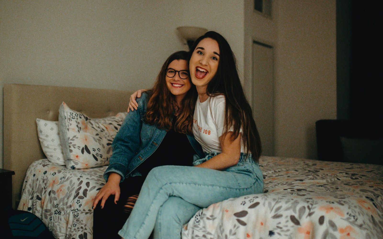 Two young women smiling and sitting together on a bed in a cozy, warmly lit Carolina Square bedroom—the perfect roommate vibe.