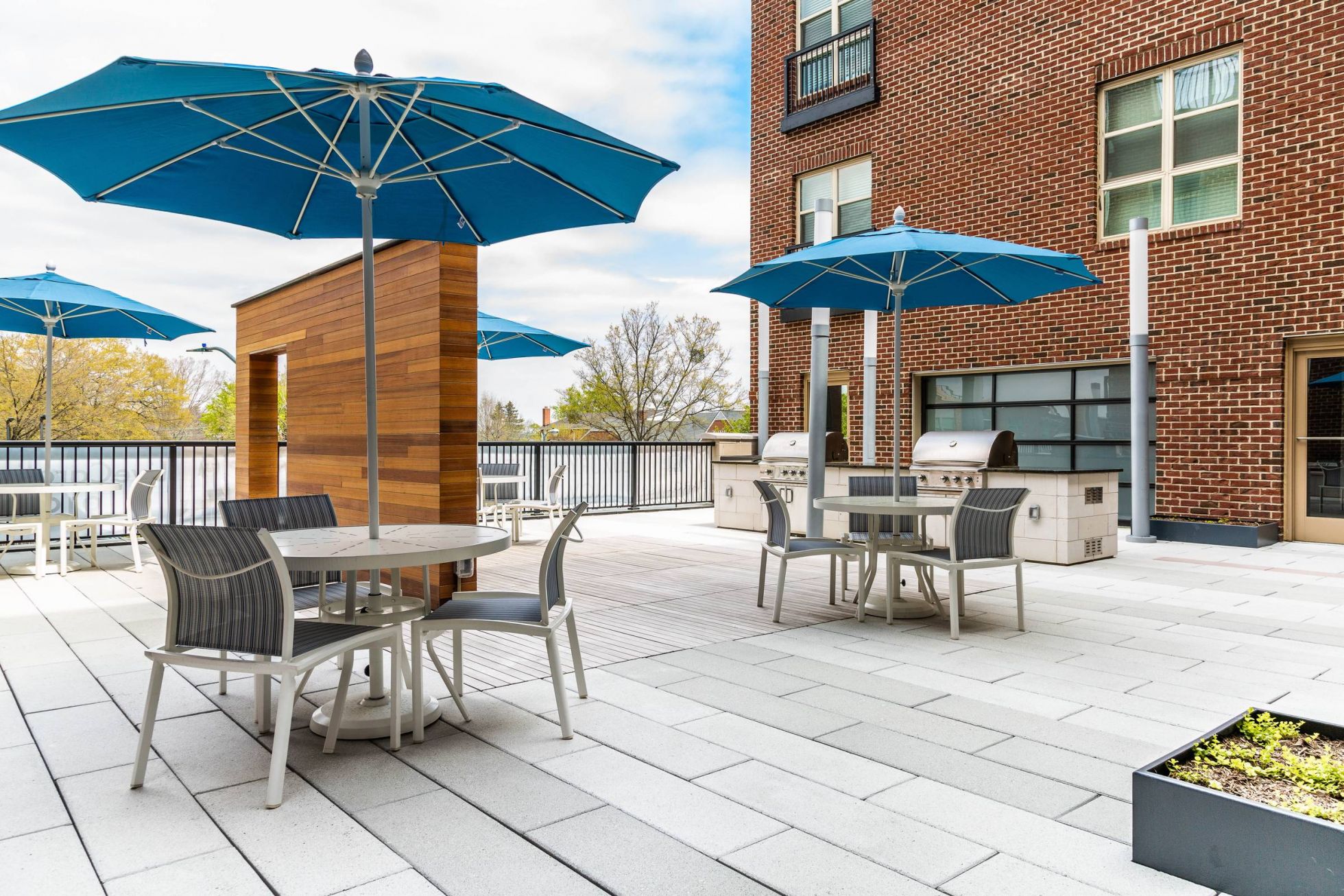 Outdoor patio with tables, chairs, blue umbrellas, and grills next to a brick building on a sunny day.