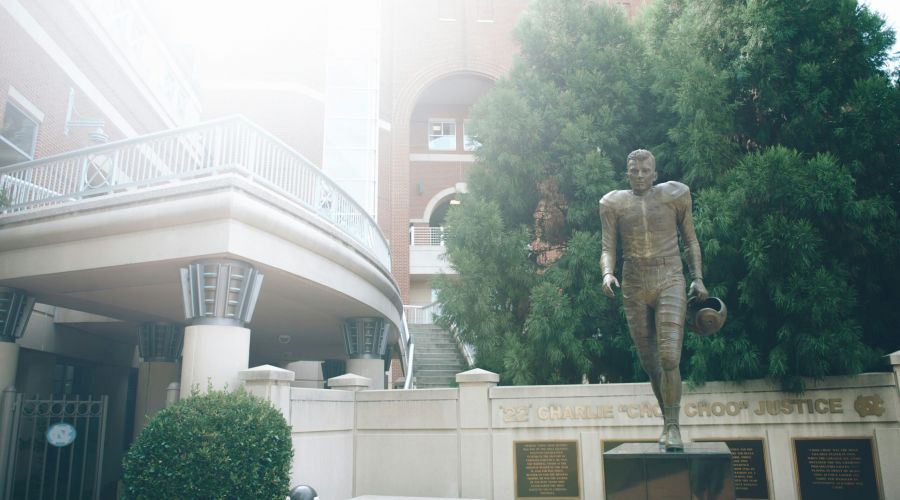 Bronze UNC football player statue holding helmet near a building and tall green tree on a sunny day.