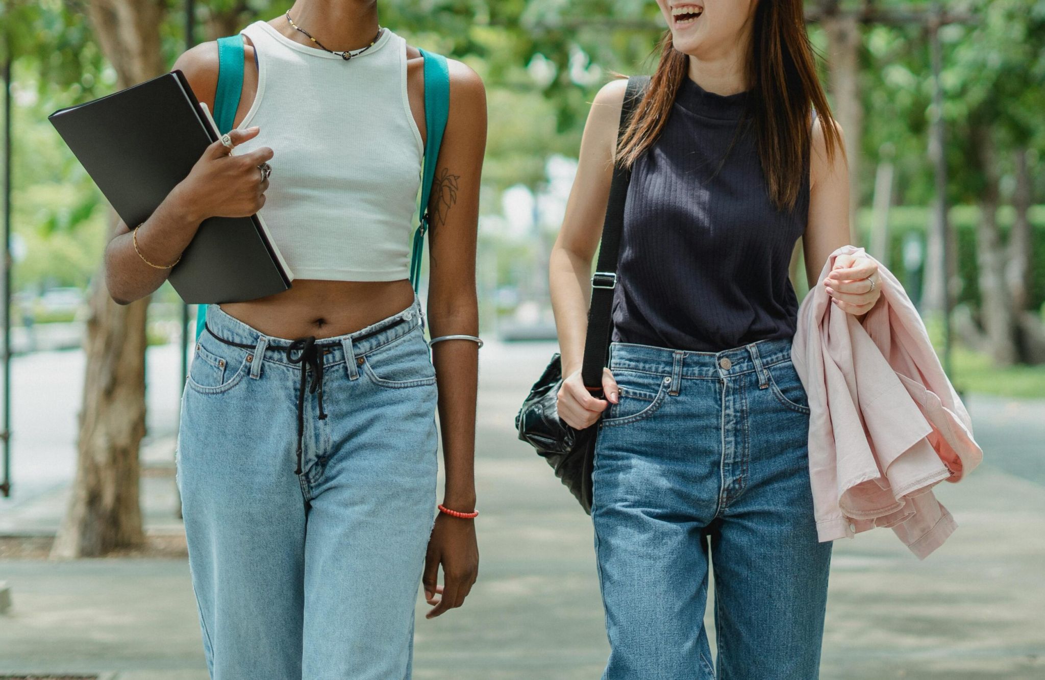 Two women walking outdoors, smiling, wearing casual jeans and sleeveless tops, one holding a laptop.