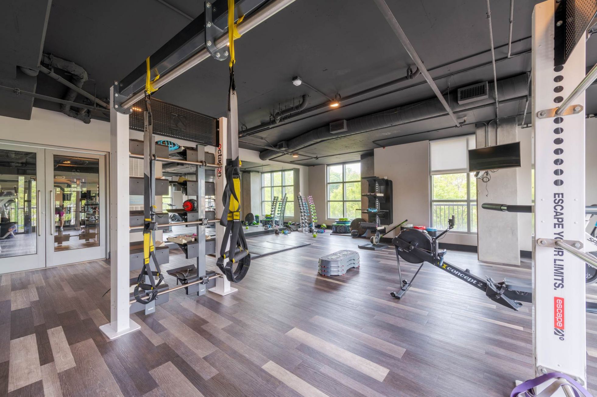 Modern gym with exercise machines, rowing machine, weights, and large windows letting in natural light.