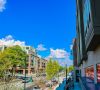 A sunny city street lined with modern buildings, trees, and parked cars under a bright blue sky.