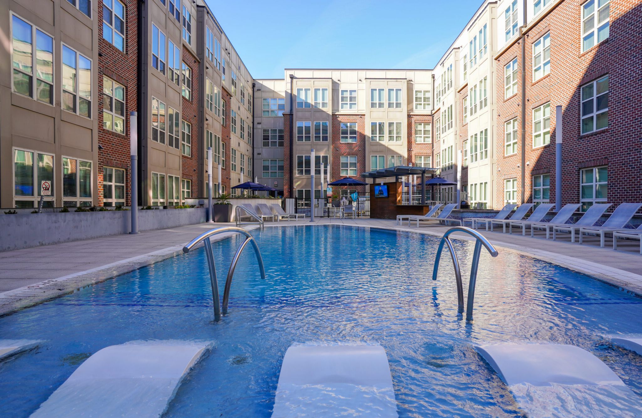 Outdoor apartment pool with lounge chairs, curved metal water features, and surrounding modern brick buildings.