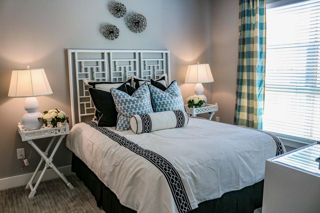 Modern bedroom with geometric headboard, patterned pillows, bedside lamps, flowers, and plaid curtains at Carolina Square apartments.