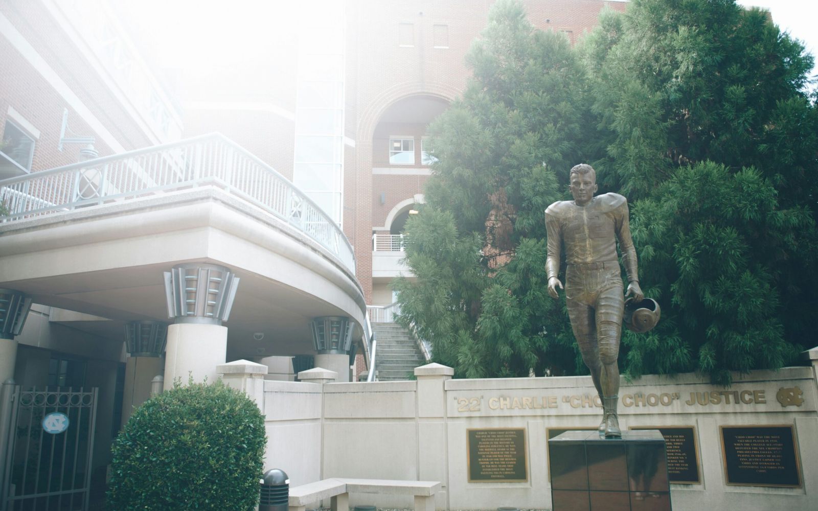 Bronze UNC football player statue holding helmet near a building and tall green tree on a sunny day.