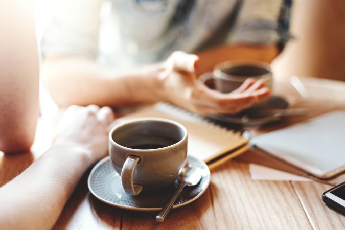 Two people talk over coffee at a wooden table with notebooks, a pen, and a smartphone.
