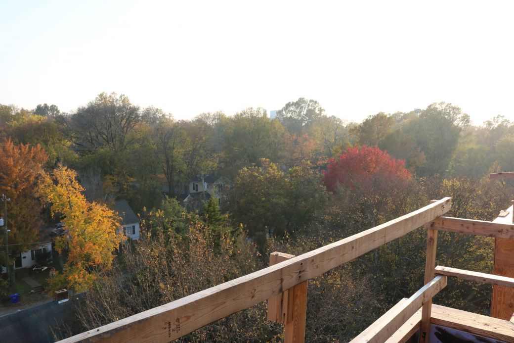 View from a wooden balcony overlooking trees with autumn foliage and houses on a sunny day.