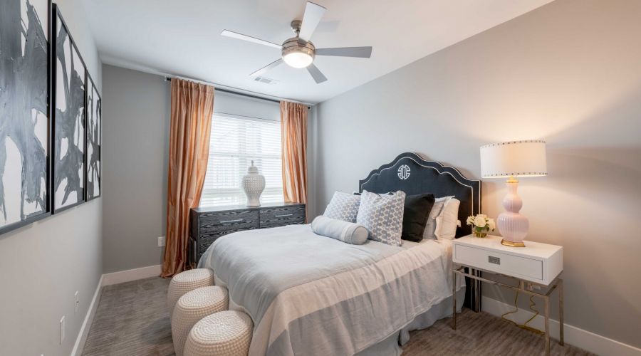 Modern bedroom with a bed, two stools, dresser, lamp, and large window with curtains letting in daylight.