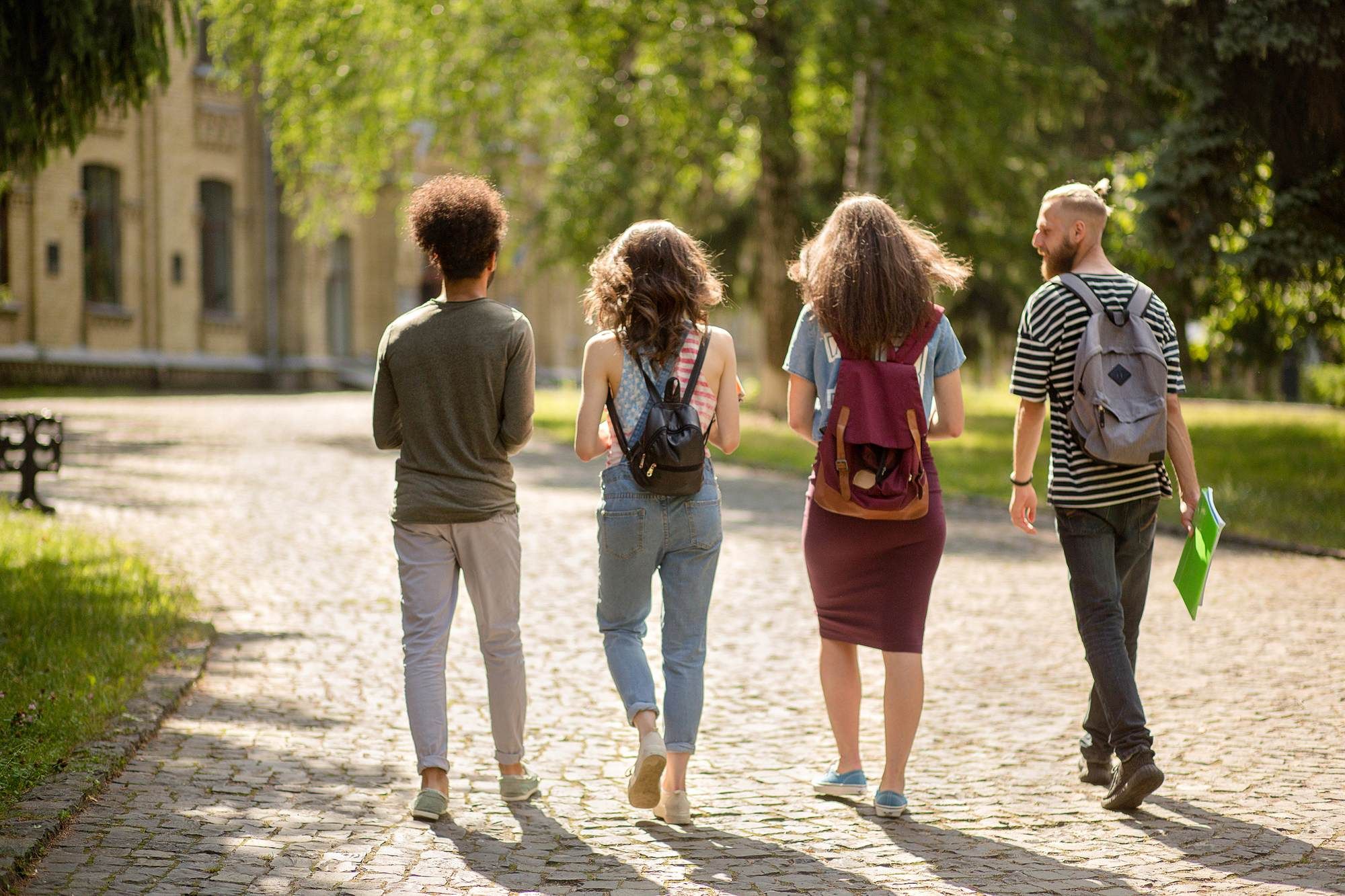 Four young adults with backpacks walk together on a sunny, tree-lined cobblestone path.