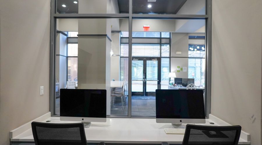 Two desktop computers on a desk in front of large windows inside a modern office space.