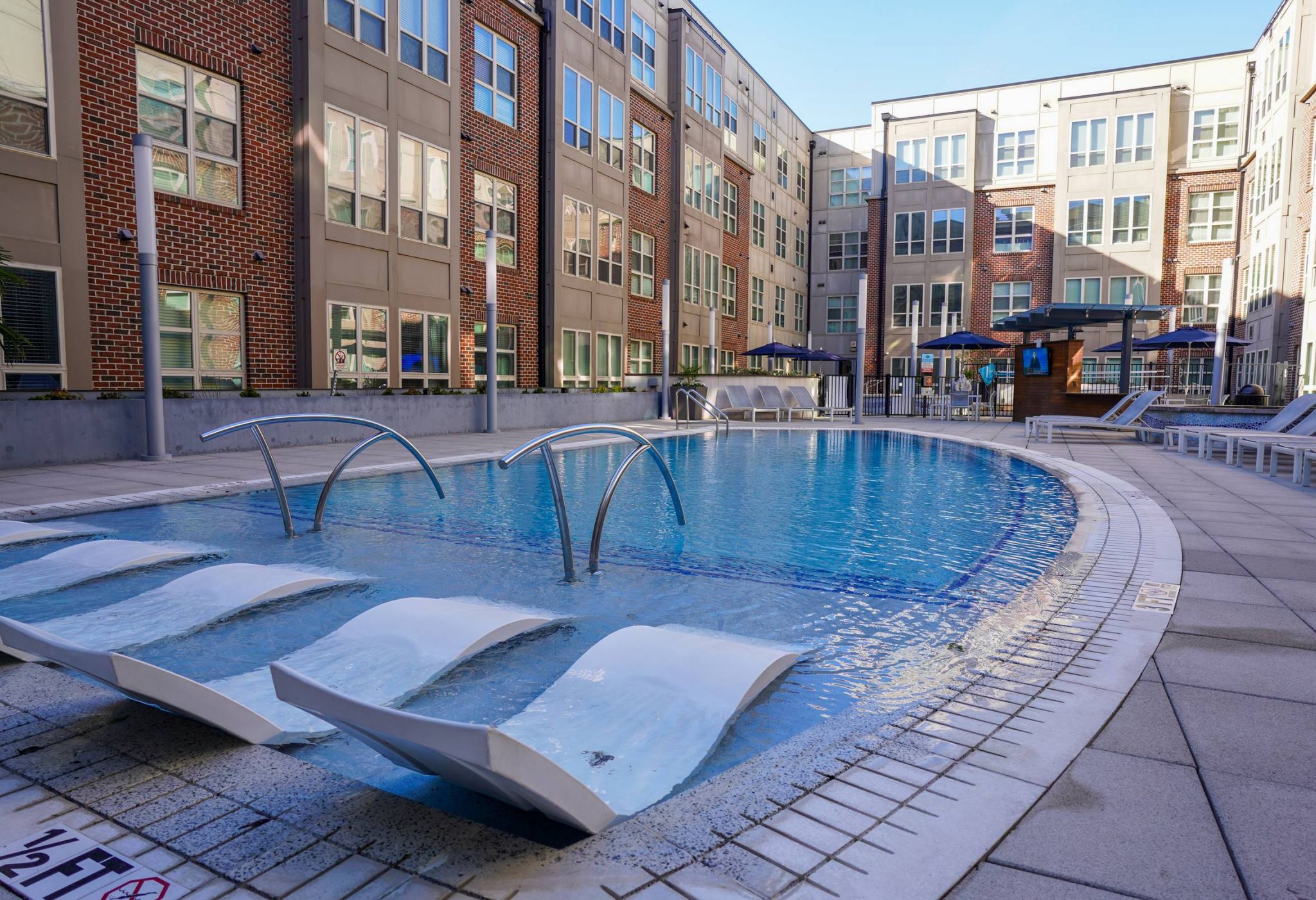 Outdoor apartment pool with lounge chairs, curved metal water features, and surrounding modern brick buildings.